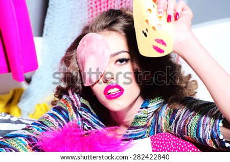 Portrait of beautiful blazing girl holding slice of cheese with pink lipstick kiss and bacon on face looking forward amid colorful clothes on grey wall background, horizontal picture