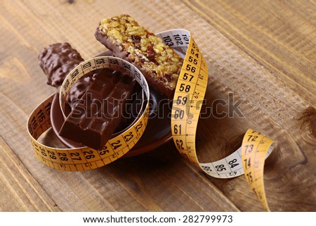 Appetizing chocolate bars and peanut brittle with a measuring tape as a symbol of diet on brown wooden table top background, horizontal photo