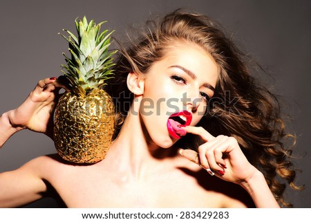 Sensual young girl with curly hair and bright pink lips holding golden pineapple on shoulder looking forward with open mouth touching her tongue with finger standing on dark grey background