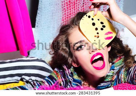 Portrait of beautiful bright girl holding slice of cheese with pink lipstick kiss looking forward surprised with open mouth amid colorful clothes on grey wall background, horizontal picture