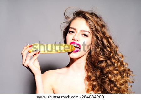 Winsome young girl with curly hair and bright pink lips holding and eating bread roll looking forward standing on grey background, horizontal picture