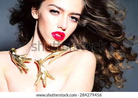 Beauteous young undressed woman with curly hair and bright pink lips with gold chicken feet as necklace looking forward standing on grey background, horizontal picture