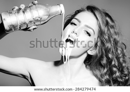 Sensual attractive young woman with curly hair holding bottle pours white yogurt on her face with open mouth looking forward standing on grey background black and white, horizontal picture