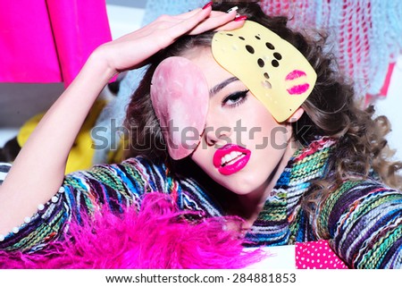 Portrait of beautiful glaring girl holding slice of cheese with pink lipstick kiss and bacon on face looking forward amid colorful clothes on grey wall background, horizontal picture