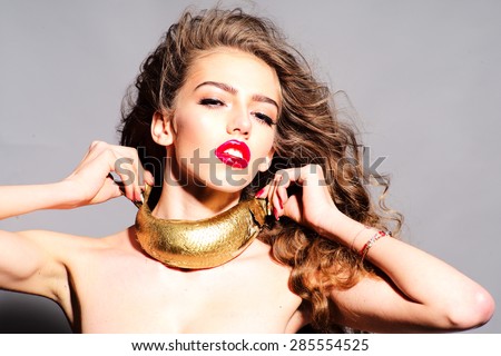Sweet young naked girl with curly hair holding golden fish as necklace looking forward standing on grey background, horizontal picture