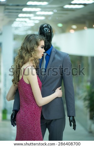 Attractive young girl with bright makeup and curly hair standing with male mannequin in formal clothes on shopping background, vertical picture