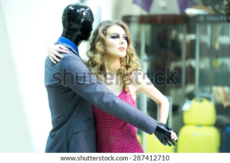 Tempting secretive young girl with bright makeup and curly hair dancing with male mannequin in formal clothes on shopping background, horizontal picture