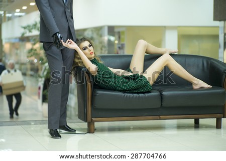 Pretty secretive young woman with bright makeup and curly hair with male mannequin lying on sofa on shopping background, horizontal picture