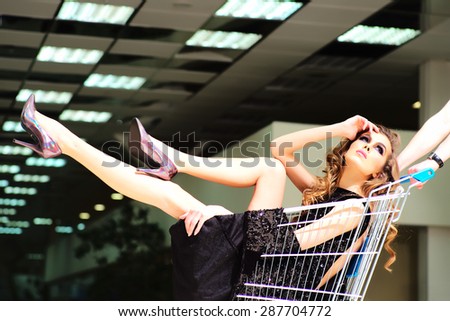 One young pretty fashion girl in black dress sitting in shopping trolley indoor on store backdrop, horizontal picture