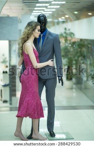Tempting young girl with bright makeup and curly hair standing with male mannequin in formal clothes on shopping background, vertical picture