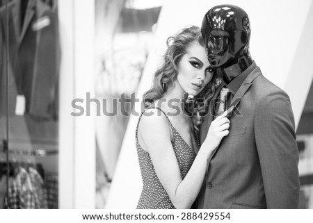 Enigmatic young woman with bright makeup and curly hair standing with male mannequin in formal clothes on shopping background black and white, horizontal picture