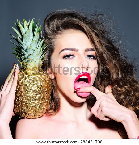 Portrait of sexy young girl with curly hair and bright pink lips  holding golden pineapple on shoulder looking forward with open mouth touching her tongue\
on dark background, square picture