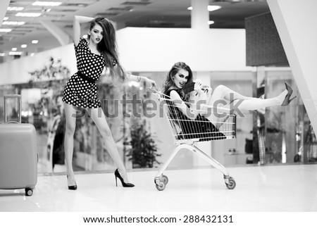 Two cute beautiful girls in dresses with shopping trolley and bag indoor on shop background black and white, horizontal picture