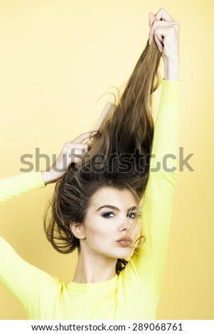 Portrait of tempting pretty young woman with long hair in yellow blouse looking forward combing hair with brush  standing on yellow background, vertical picture