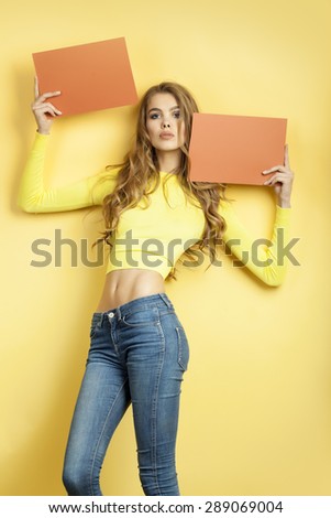 Slender beautiful young woman with long curly hair in yellow blouse and blue jeans holding two orange brown sheets of paper standing on yellow background, vertical picture