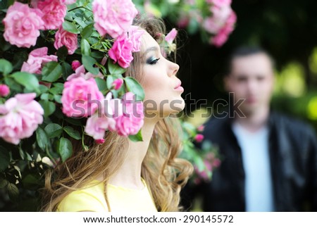 Sensual young blonde lady with bright makeup and curly hair in yellow dress smelling lush bush of pink rose and man in background, horizontal picture