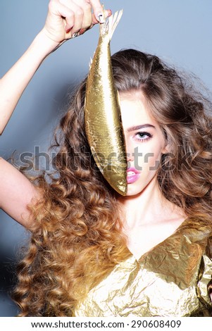 Winning young girl with curly hair in gold jacket holding golden fish covering half face looking forward standing on grey background, vertical picture