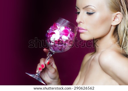 Young beautiful tender girl smelling pink purple and white flower petals in wine glass standing in studio on dark violet background copyspace, horizontal picture