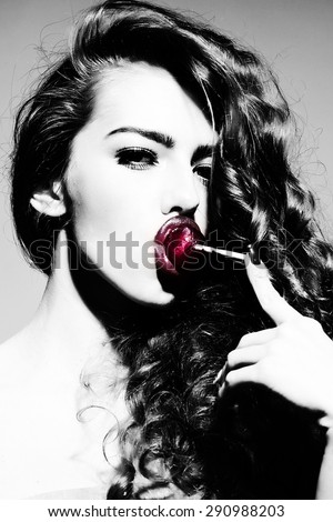 Portrait of fascinating playful young undressed woman with curly hair and bright pink lips holding with finger purple round lollipop in mouth looking forward standing black and white, vertical picture