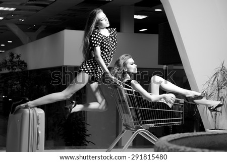 Two alluring fashionable girls in dresses with shopping trolley indoor on shop background black and white, horizontal picture