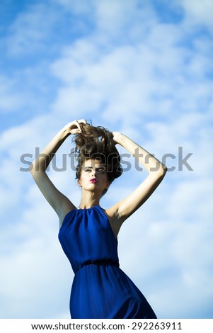 Young pretty sexual woman in long blue dress with curly hair standing on light blue sky and white clouds background, vertical picture