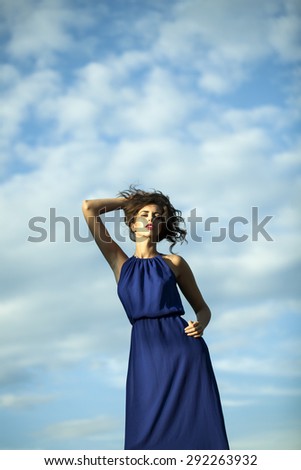Young beautiful sexy woman in long blue dress with curly hair standing on light blue sky and white clouds background, vertical picture