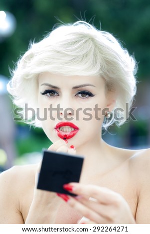Portrait od pretty young sexy blonde lady with red nails put on lipstick holding small mirror standing on natural background closeup, vertical picture