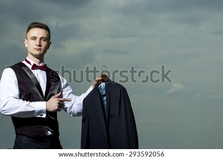 Handsome sexual young strong muscular man in formal suit with bow tie holding jacket standing on blue sky background copyspace, horizontal picture