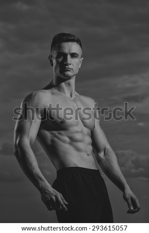 Beautiful sexy muscular naked beefy male bodybuilder standing on sky background black and white, vertical picture