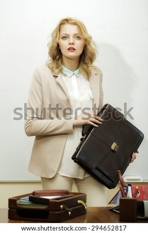 Solid beautiful bussiness woman in formal suit holding dark brown leather case standing near table with office supplies on white background, vertical picture