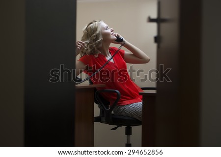 Pretty sexual blonde woman secretary with curly hair in red blouse sitting in private cabinet at table speaking by phone view from door, horizontal picture