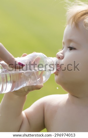 Closeup of little male child with blonde curly hair round cheeks and cute face drinking water from plastic bottle from hand of mother sunny day outdoor on green grass background, vertical picture