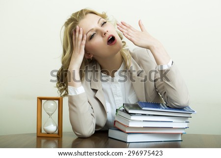 Portrait of yawning pretty young woman with blonde curly hair in jacket sitting at table with heap of books and sand hour glass clock on white wall background, horizontal picture