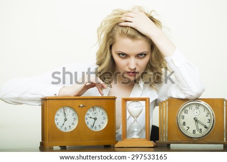 Attractive serious waiting young blonde business woman sitting in shirt with wooden sand glass and clock with dial looking forward on white background, horizontal picture