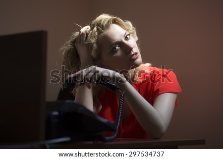 Thoughtful beautiful secretary blond woman sitting at office table underlaying in red blouse holding phone receiver on bare wall background, horizontal picture