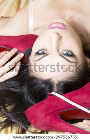 Sexy beautiful young brunette woman model with bright makeup and soft lips lying looking forward and holding pair of two fashionable red suede leather shoes on high heels, vertical picture