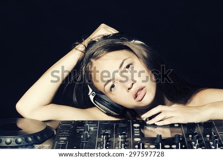 Portrait of young pretty sexual disk jockey girl in headphones looking forward lying on professional musical mixer console on black studio background, horizontal picture