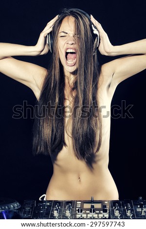 Beautiful sexy undressed young disc jockey woman standing near mixer console shouting and holding head phones with hands on black background, vertical picture