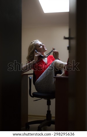 View from doorway of smiling beautiful blonde secretary woman sitting in office with feet on table in red blouse holding telephone receiver speaking on phone, vertical picture