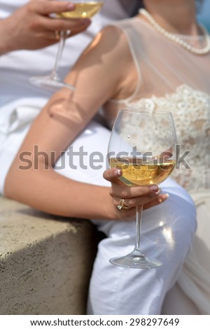 Young wedding couple of two people in white clothes holding glasses with white wine sitting and standing sunny day outdoor, vertical picture