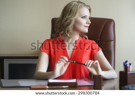 Pretty smiling elegant blond business woman sitting in office on brown leather chair in red blouse holding pen in hands looking away indoor on white background, horizontal picture
