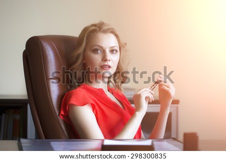 Beautiful young elegant blond business woman sitting in office on brown leather chair in red blouse holding pen in hands looking forward indoor on white background copyspace, horizontal picture
