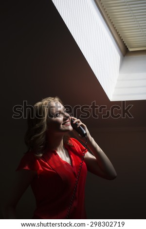 Beautiful smiling young blonde woman with curly hair in red red blouse standing in office looking at window and speaking on phone indoor, vertical picture
