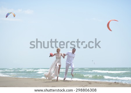 Beautiful laughing young wedding couple of man and woman in white jumping and running along ocean beach shore on windy weather sunny day with paraplanes on blue sky background, horizontal picture