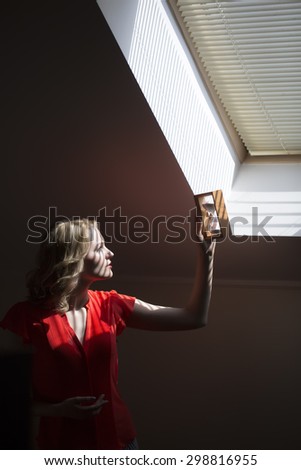 Attractive pensive young blonde woman with curly hair in red red blouse standing in office near window holding clock and looking on time at sand glass indoor, vertical picture