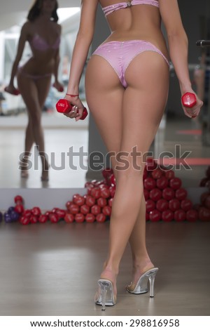 Back view of beautiful sexy fitness body of young sporty woman with straight legs on high heels in erotic pink bikini with sparkles standing with red dumb bells reflecting in mirror, vertical picture