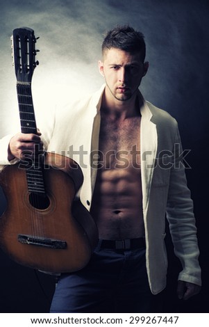 Handsome sexy young musical macho man with muscular body in jeans and white jacket with naked torso holding wooden acoustic guitar looking forward standing on grey glare background, vertical picture