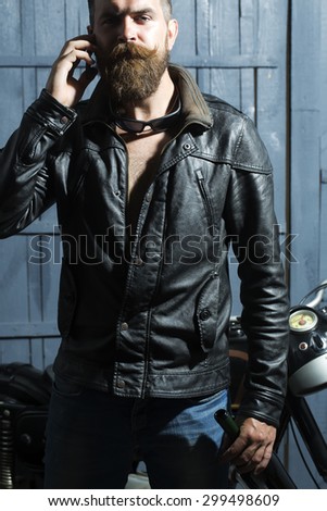 Handsome brutal unshaven sexy male biker in brown leather jacket blue jeans and sun glasses standing in garage near motor cycle holding mobile phone and speaking on wooden background, vertical picture