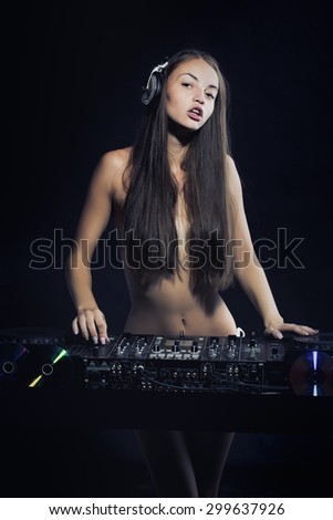 Beautiful naked musical dj young girl with long hair standing near mixer console with head phones on black background, vertical picture