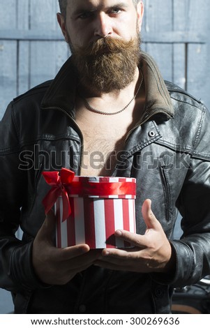 Handsome unshaven young guy with beard and moustache in brown leather jacket and chain holding round red white striped present box with ribbon bow standing on grey wooden background, vertical picture